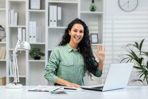 Portrait of young beautiful business woman at workplace inside office, Hispanic woman smiling and looking at camera, waving hand greeting gesture, working with laptop remotely in home light office. photo