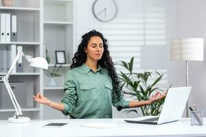 Young beautiful hispanic woman relaxing in home office, successful businesswoman meditating in lotus position sitting at workplace with laptop, freelancer in green shirt in living room. photo