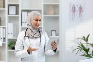 Young experienced female doctor in hijab using tablet computer for video call, female clinic worker consults patients remotely online, smiling contentedly inside clinic office. photo