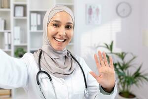 hermosa asiático musulmán hembra médico en hijab y traje tomando selfie foto mientras descansando un médico hace un vídeo llamar, sonriente y ondulación como él saluda su familia desde su lugar de trabajo