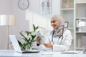 retrato musulmán hembra médico en hiyab, mujer médico con tableta computadora utilizando solicitud, sonriente satisfecho trabajo trabajando dentro médico oficina, en blanco médico abrigo, sentado a mesa con ordenador portátil foto