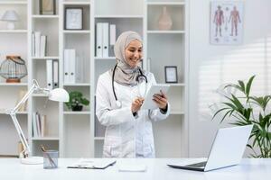 joven hermosa hembra médico en hijab utilizando tableta computadora, médico trabajador en blanco médico Saco y estetoscopio dentro clínica oficina a lugar de trabajo, sonriente musulmán mujer utilizando en línea solicitud. foto