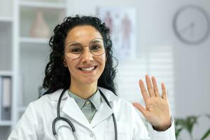 Online video consultation, female doctor in white medical coat talking on video call smiling and looking at camera, Latin American woman with stethoscope waving joyfully greeting gesture. photo