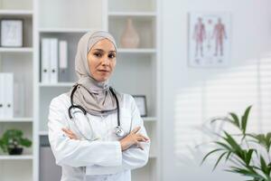 retrato de grave confidente exitoso hembra médico en hijab en blanco médico Saco mirando a cámara con cruzado brazos, clínica trabajador trabajando dentro oficina médico habitación. foto