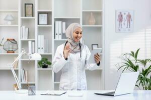 un sonriente musulmán médico en un hijab conduce un en línea cita con un paciente, sostiene un tableta, olas a el cámara y dice Hola. foto