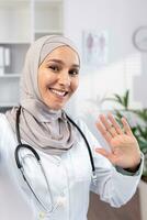 un musulmán hembra médico en un hijab conduce un en línea cita con pacientes en el teléfono, olas a el cámara, ver desde el teléfono. foto