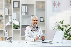 Portrait of Muslim female doctor in hijab, female doctor with tablet computer looking at camera with smile, working inside medical office, wearing white medical coat, sitting at table with laptop. photo