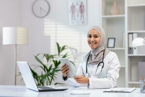 retrato de musulmán hembra médico en hiyab, hembra médico con tableta computadora mirando a cámara con sonrisa, trabajando dentro médico oficina, vistiendo blanco médico abrigo, sentado a mesa con ordenador portátil. foto