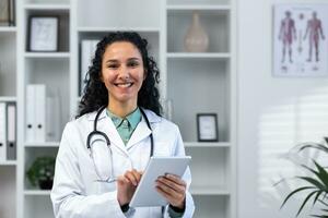 retrato de exitoso sonriente Hispano hembra médico dentro médico oficina, mujer con tableta computadora cerca arriba sonriente y mirando a cámara, trabajando en médico vestido mientras de pie. foto