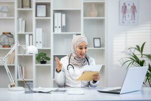 Joyful female doctor in hijab received envelope message mail notification with good results of patient's treatment, woman is happy and smiling, working inside clinic office in white medical coat. photo