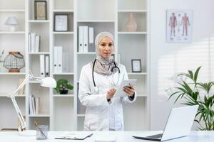 Closeup portrait of Arabic female doctor in hijab and white medical coat standing with tablet in hands in private doctor's office, looking at camera. photo