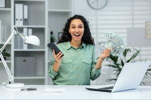 Online casino. Young muslim woman sitting in office at table with laptop, holding cash in hand and phone, playing online games, making bets. Smiling at the camera, celebrating the win. photo