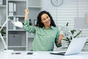 A young Hispanic woman is sitting at the desk in the office and holding money in her hands. Happy and smiling. Celebrates, dances. photo