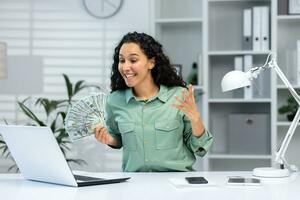 exitoso mujer de negocios a lugar de trabajo dentro oficina participación efectivo dinero dolares en manos, celebrando alegre alto logro resultados, trabajando con ordenador portátil. foto