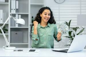 exitoso mujer de negocios a lugar de trabajo dentro oficina participación efectivo dinero dolares en manos, celebrando alegre alto logro resultados, trabajando con ordenador portátil. foto