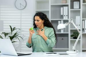 Woman with sore throat at workplace inside office, business woman with laptop working at work, Hispanic woman using medicine to relieve sore throat. photo