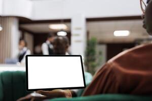 Female guest holding device at reception, relaxing in lounge area with digital tablet and blank screen template. Woman using white display with isolated copyspace at front desk lobby. photo