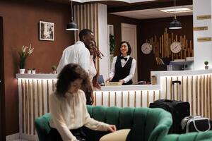 Happy couple arriving in hotel lobby and preparing to check in for room reservation at reception desk. Young guests registering on booking accommodation, staff welcoming people. photo