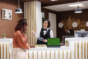 Diverse women using greenscreen display at hotel front desk, talking about check in process and registration. Female guest looking at isolated copyspace template with blank chroma key. photo