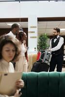 Male employee helping clients with bags, carrying luggage for people after doing check in procedure. Young couple receiving assistance from luxury resort bellboy at reception desk. photo