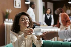 Hotel porter serving woman with coffee, giving beverage to hotel manager at reception. Administrator sitting in lounge area at resort, receiving drink from bellboy with professionalism. photo