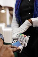 Man making payment using contactless credit card, closeup of hand paying bill order with NFC technology while resting at hotel lounge area. Waitress wearing white gloves holding POS terminal photo