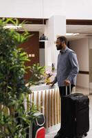 Young African American man with suitcase standing at reception area, talking pleasantly with front desk employees, waiting in line to check-in after arrival. Black guy tourist checking out of hotel photo