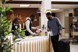 Front desk staff checking guest passport and travel documents during hotel check-in procedure. African American man receptionist using computer software, tourist with suitcase at reception photo