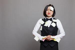 Unhappy exhausted asian waitress covered with tasks sticky notes standing portrait. Depressed overloaded woman receptionist in uniform looking at camera with upset facial expression photo