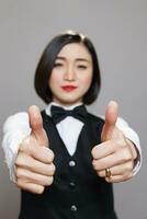 Cheerful asian woman in waitress uniform smiling and giving thumbs up symbol closeup. Cafe worker confirming and recommending restaurant good service and looking at camera photo