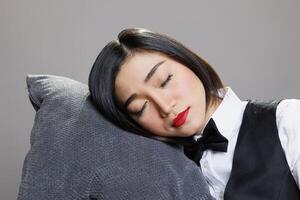 agotado soñoliento asiático camarera descansando cabeza en almohada, posando en estudio con cerrado ojos. joven atractivo cansado restaurante recepcionista vistiendo uniforme dormido de cerca foto