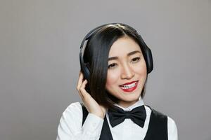 Smiling asian waitress in uniform listening to music in wireless headphones. Cheerful young attractive restaurant receptionist wearing earphones and enjoying playlist while posing in studio photo