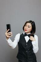 Smiling woman receptionist chatting in remote conversation on mobile phone. Young attractive asian waitress looking at smartphone front camera while taking selfie and posing in studio photo