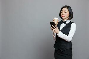 Young asian waitress wearing uniform drinking coffee to go and scrolling social networks on smartphone. Attractive woman receptionist holding paper mug and using mobile phone photo