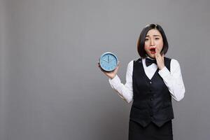 Hotel asian woman receptionist yawning while holding alarm clock and looking at camera in studio. Young attractive tired sleepy waitress oversleeping and showing time portrait photo