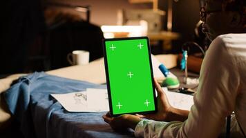 Designer using greenscreen on tablet, looking at modern chroma key display in atelier workshop. Couturier holding device with isolated mockup on screen, custom made tailoring. Close up. photo