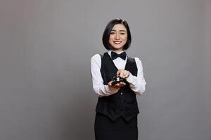 Smiling asian restaurant receptionist in uniform ringing dining bell while posing and looking at camera. Young waitress pressing order notification alert portrait on gray background photo