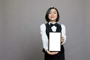 Smiling young waitress in professional uniform displaying digital tablet empty screen with copy space. Asian receptionist presenting white touchscreen for catering service application advertising photo