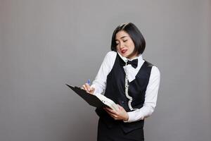 Waitress holding clipboard and talking on landline phone, managing communication and tasks in cafe. Receptionist engaging in conversation on telephone and writing information photo