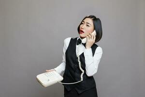 asiático mujer recepcionista en uniforme hablando en teléfono fijo teléfono, dando instrucción a restaurante personal. confidente restaurante trabajador respondiendo cliente llamada utilizando retro teléfono en estudio foto