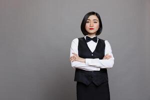 Catering service young asian waitress posing with crossed arms showcasing professionalism and looking at camera. Restaurant receptionist wearing black and white uniform portrait photo