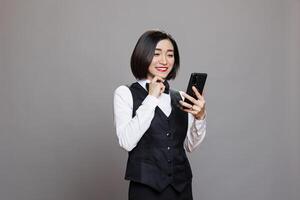 Happy asian cafe waitress reading client positive feedback on smartphone. Cheerful smiling receptionist in professional uniform checking customer satisfaction while using mobile phone photo