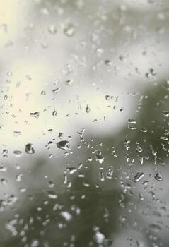 A photo of rain drops on the window glass with a blurred view of the blossoming green trees. Abstract image showing cloudy and rainy weather conditions
