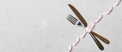 Top view of crossed knife and fork connected by measuring tape on cement background. Overeating and healthy diet concept photo
