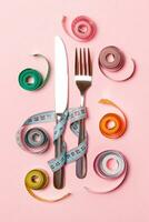 Composition of fork and knife surrounded with colored balled measuring tapes on pink background. Top view of obesity concept with copy space photo