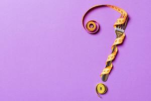 Top view of fork and measure tape in form of question mark on purple background. Overeating concept and copy space photo