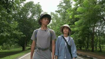 Two Asian young teen explorers with backpack walking along together on the road in nature park during summer camping. video