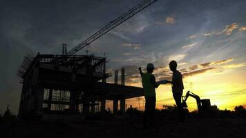 Silhouettes of architect civil and engineer in protective helmets and vests are shaking hands while working in construction site. video