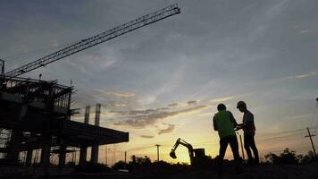 Silhouettes of architect civil and engineer in protective helmets and vests are shaking hands while working in construction site. video