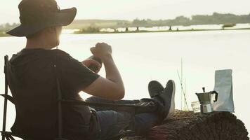 silhouet van jong Mens wie zit in een vouwen stoel Aan rivier- aan het kijken zon opstaan. drinken koffie, koffie grond, reizen levensstijl succes concept avontuur vakanties buitenshuis vrijheid emoties. video